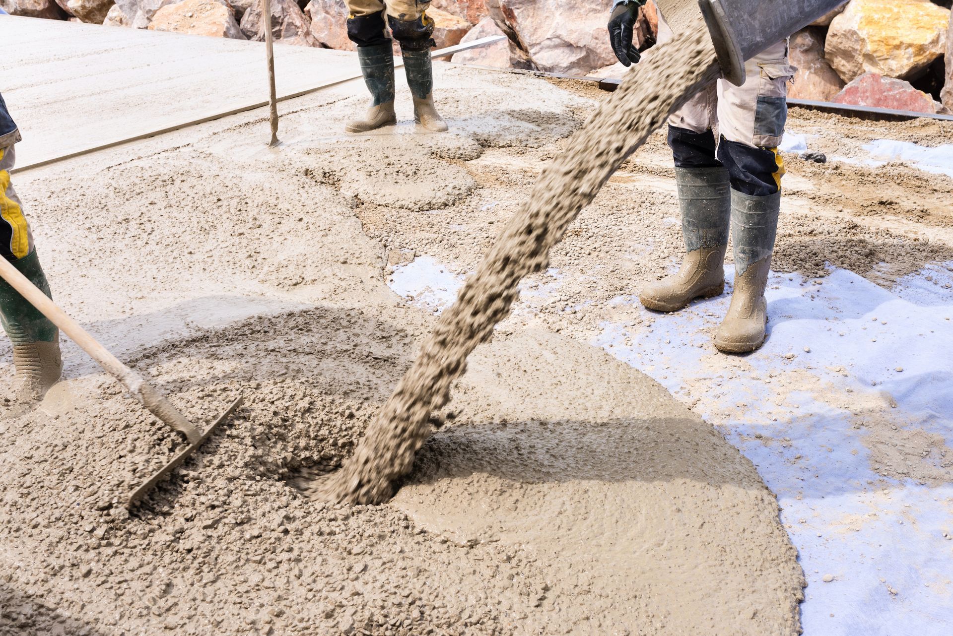 Du béton est déversé d'une goulotte sur le sol tandis que des ouvriers utilisent un râteau pour niveler et étaler le matériau humide.