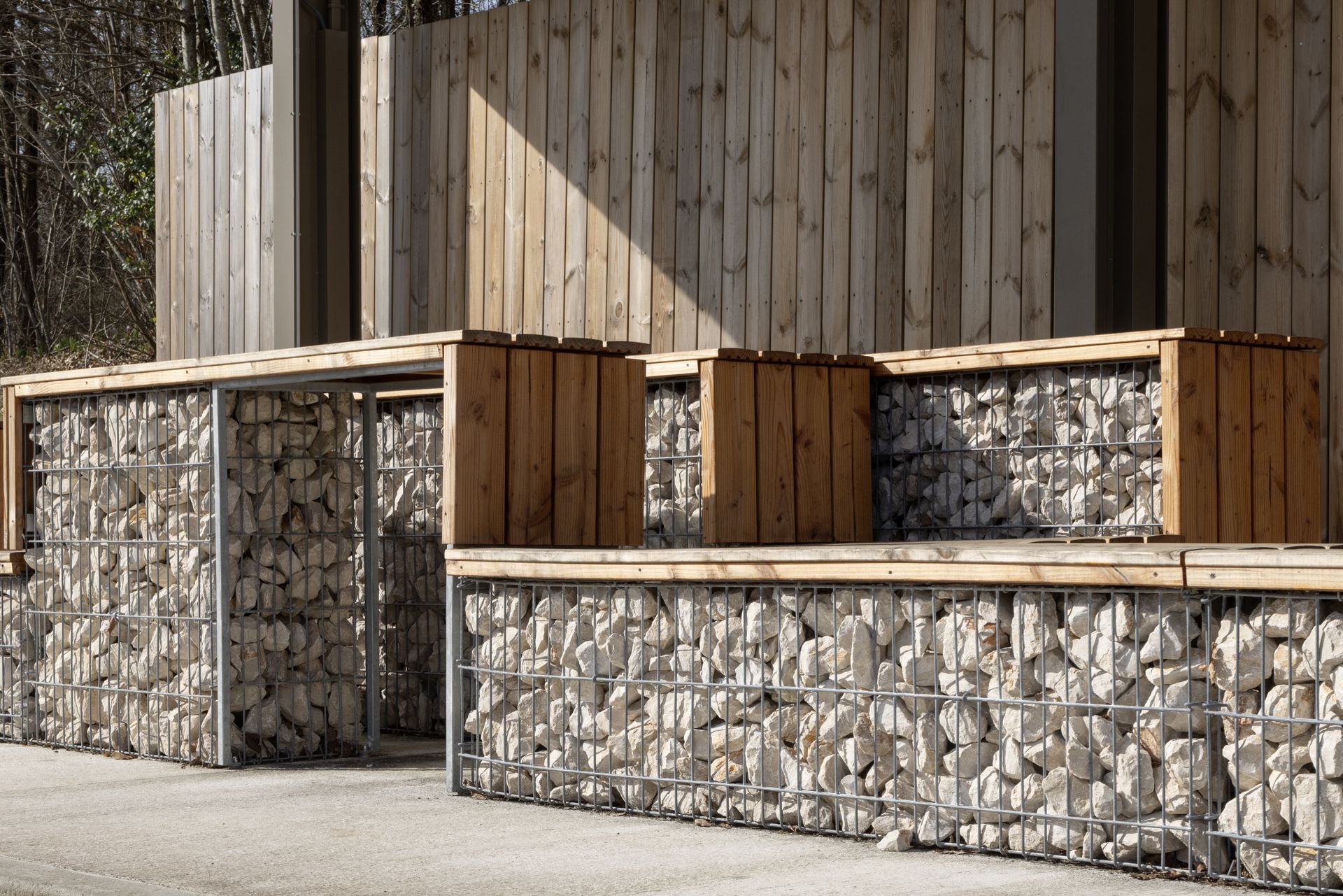Des sièges d'extérieur modernes composés de gabions en fil de fer remplis de pierres blanches et recouverts de planches de bois clair.
