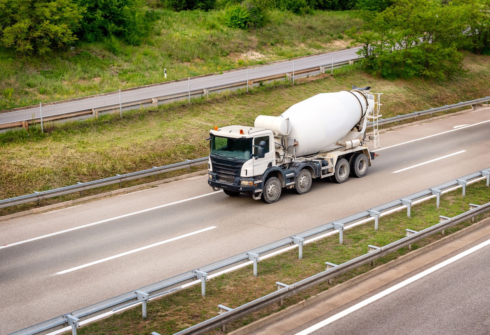 Un camion-malaxeur à ciment blanc circule sur une autoroute à plusieurs voies, entouré de collines verdoyantes et de glissières de sécurité métalliques.