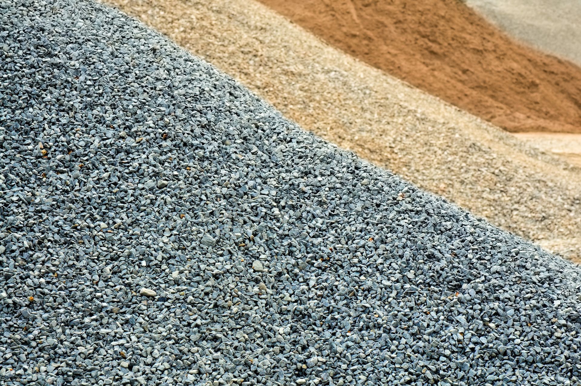 Trois grands tas contigus de matériaux de construction composés de pierres concassées grises, de gravier beige et de sable brun.