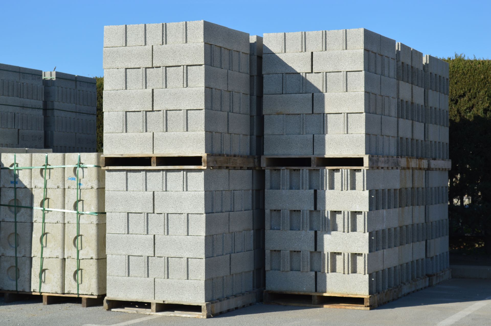 Des piles de blocs de béton gris disposées sur des palettes en bois dans une cour extérieure sous un ciel bleu azur.