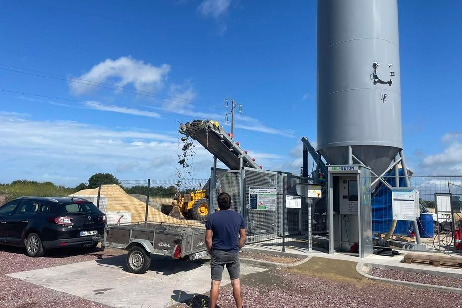 Par une journée ensoleillée, une personne se tient devant un grand silo industriel gris et un tapis roulant déposant des matériaux sur un tas.
