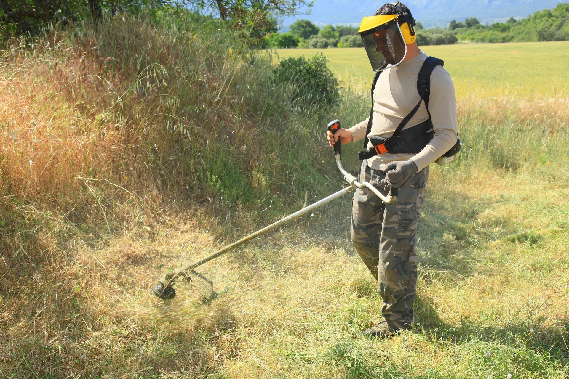 Un ouvrier utilise une débroussailleuse dans un champ avec de l'herbe envahi de hautes herbes, sous un soleil éclatant.