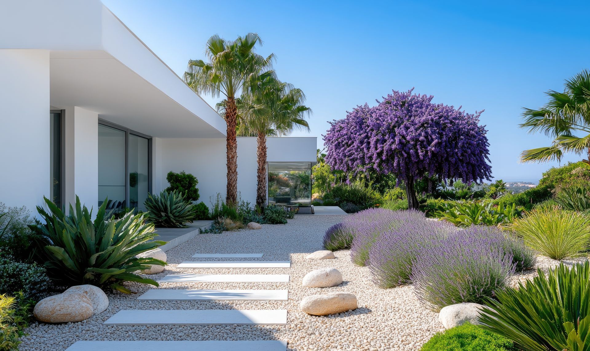 Jardin moderne jouxtant une maison blanche, avec allées de gravier, palmiers, arbustes de lavande et un arbre pourpre.
