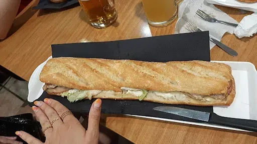 Un sándwich largo con lechuga, carne y salsa sobre una pizarra negra, sobre una mesa con bebidas y cubiertos.