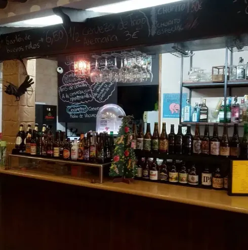 Barra de bar con varias botellas de cerveza y licor; menú en pizarra encima.