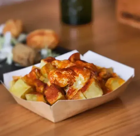 Patatas bravas en un recipiente de papel, servidas en una mesa de madera. Patatas bañadas en salsa picante.