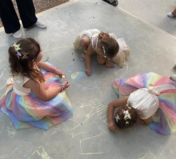 Trois enfants en robes colorées dessinent à la craie sur du béton.