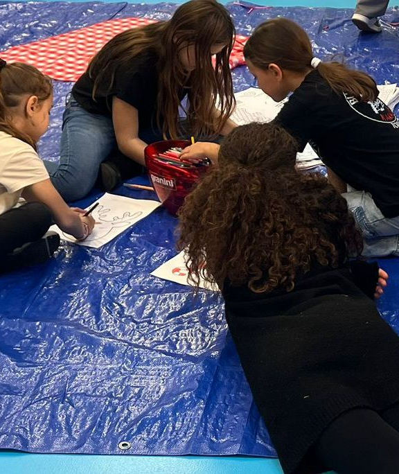 Des enfants dessinent sur du papier étalé sur une bâche bleue.
