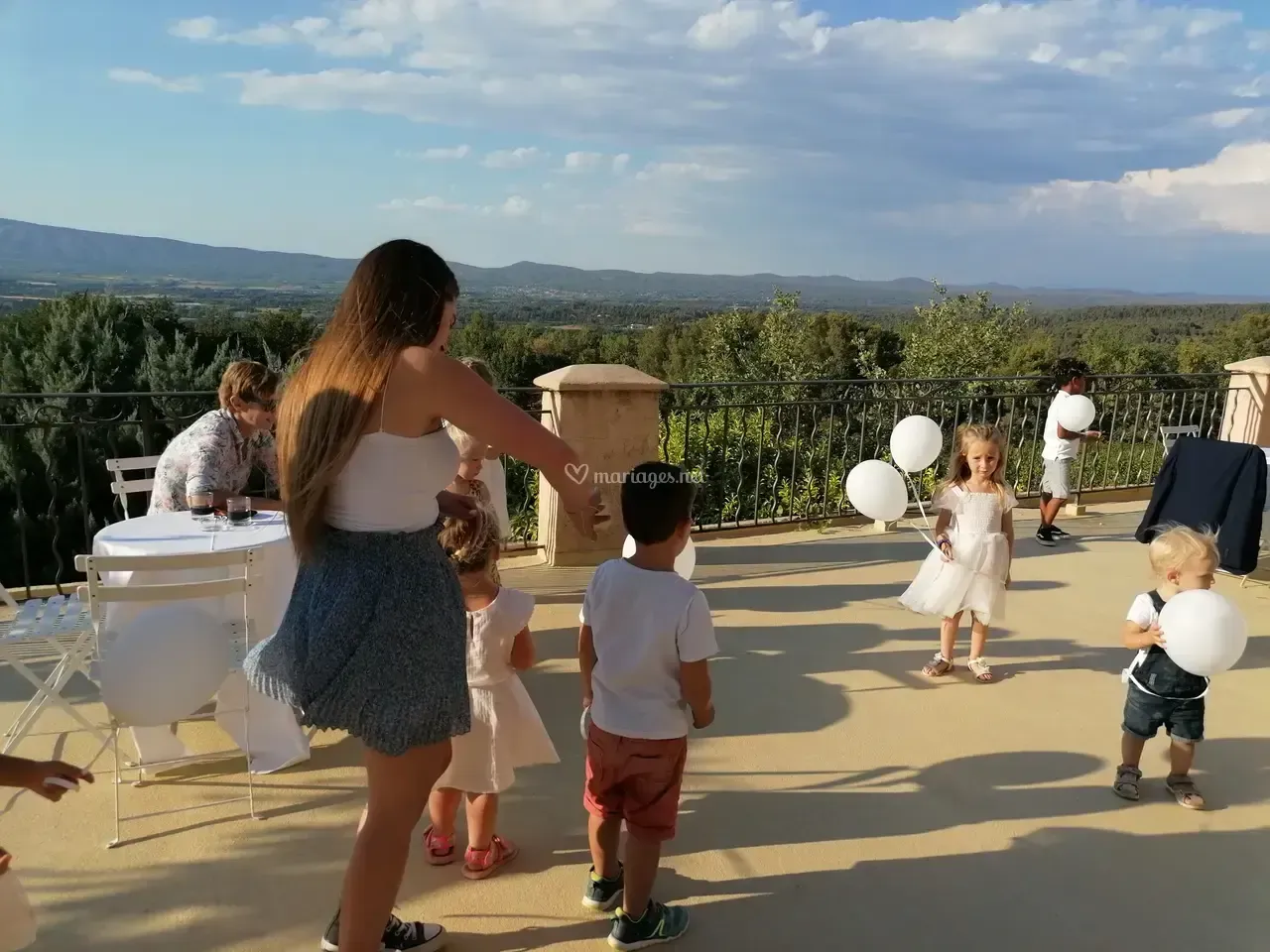 Des enfants jouent sur une terrasse avec des ballons blancs, des adultes en arrière-plan.