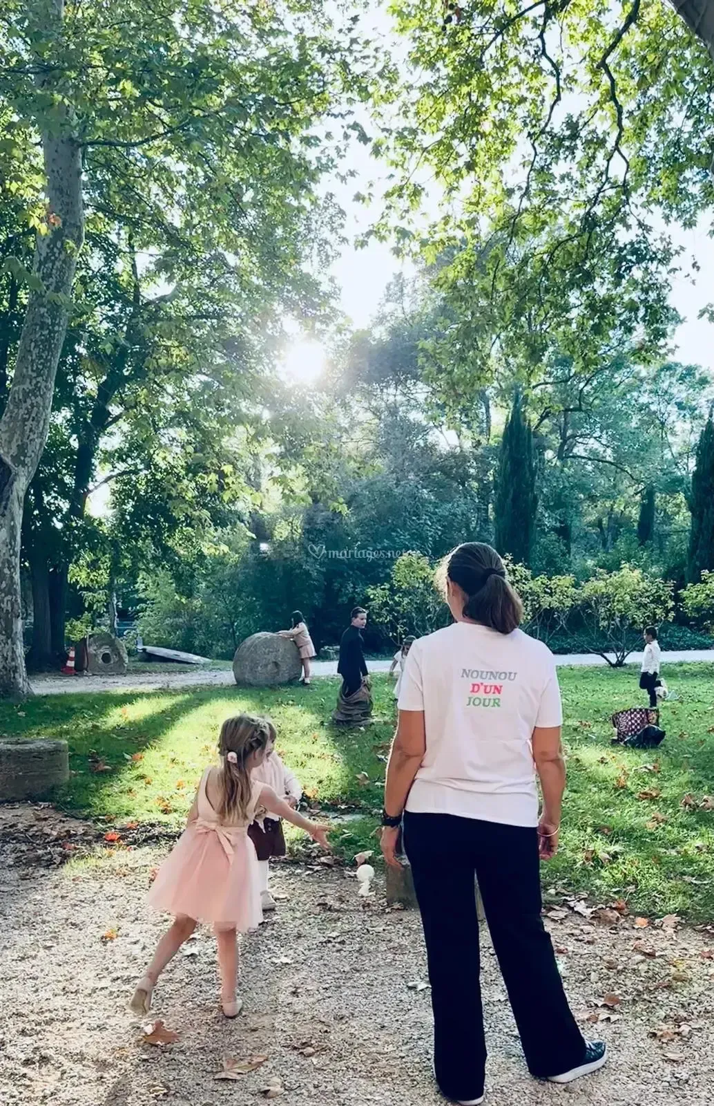 Une enfant en robe rose montre du doigt les arbres ; un adulte en chemise blanche observe la scène dans un parc ensoleillé.