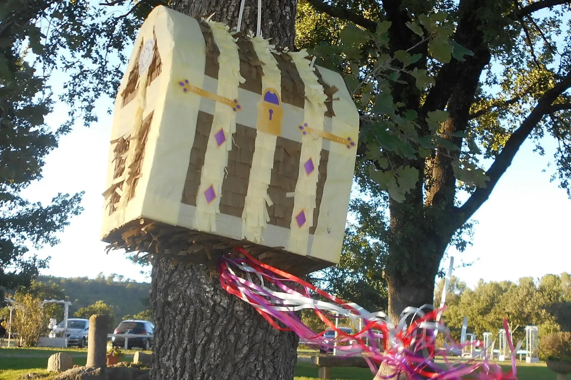 Une piñata en forme de coffre au trésor, ornée de rayures brunes, est suspendue à un arbre.