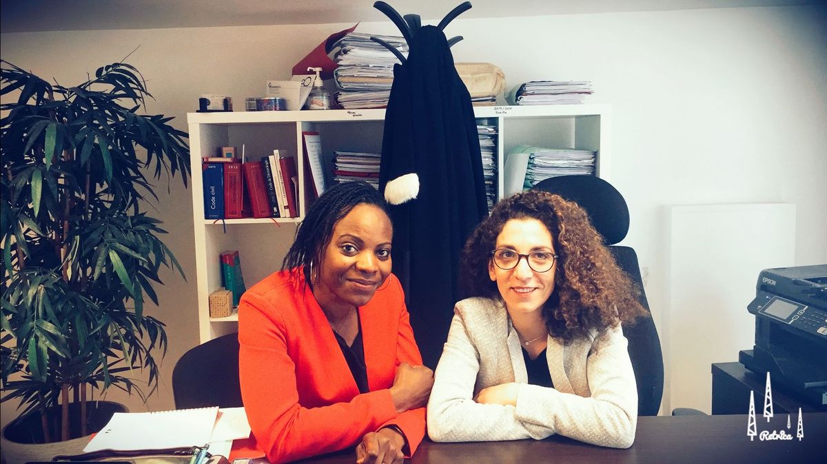 Deux collègues sourient à un bureau dans un bureau, avec des étagères et un porte-manteau en arrière-plan.