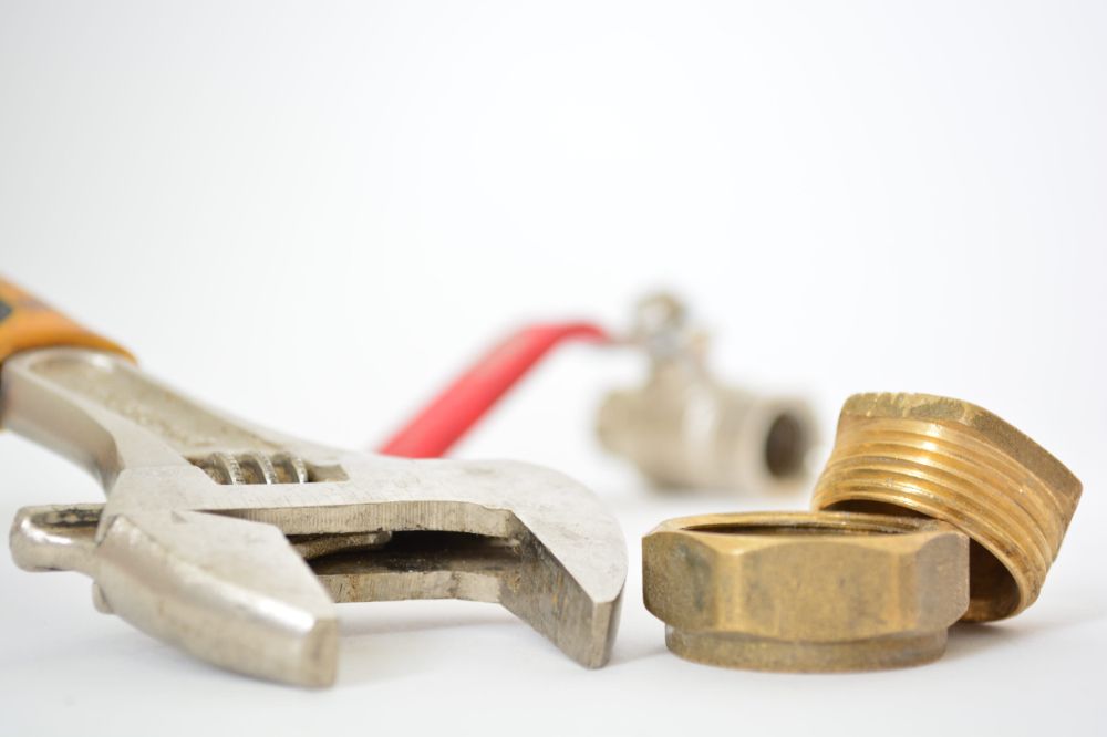 Una llave inglesa ajustable yace sobre una superficie blanca junto a un racor de tubería de latón y una válvula de bola