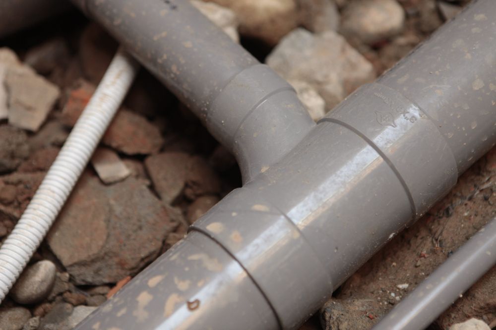 Vista de cerca de tuberías de PVC grises unidas por un conector en forma de T, que yacen sobre una capa de grava y tierra.
