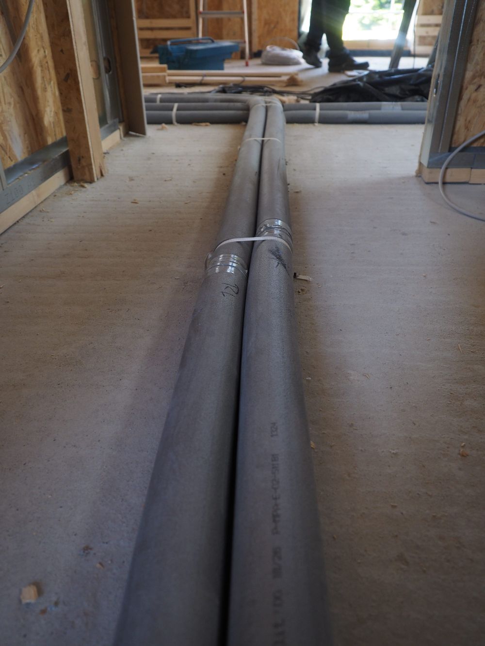 Dos tuberías grises y aisladas que atraviesan el suelo de una habitación en construcción, con un trabajador visible al fondo.