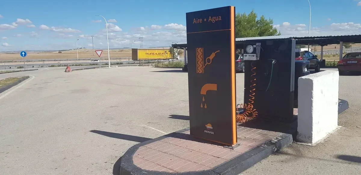 Estación de carga para vehículos eléctricos al aire libre con cargador negro y naranja junto a un área de estacionamiento bajo un cielo despejado.