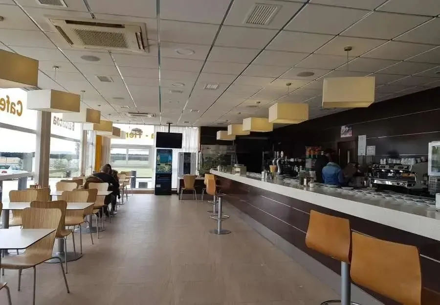Zona de estar de cafetería moderna y vacía con una larga barra y mesas bajo luces cálidas del techo.
