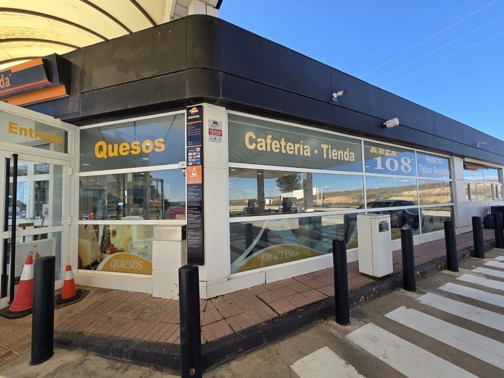 Cafetería y tienda de quesos a pie de calle con grandes ventanales, toldo negro y paso de peatones enfrente.