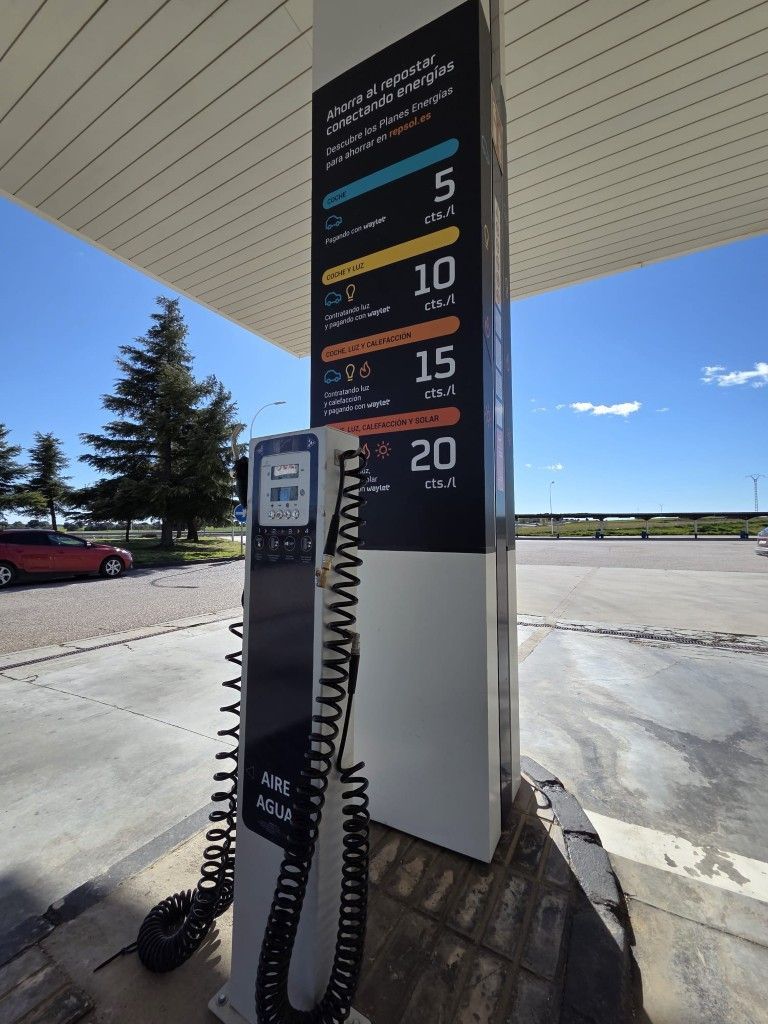 Cargador para vehículos eléctricos en una gasolinera, con cables negros colgando y un cartel con los precios 5, 10, 15 y 20.