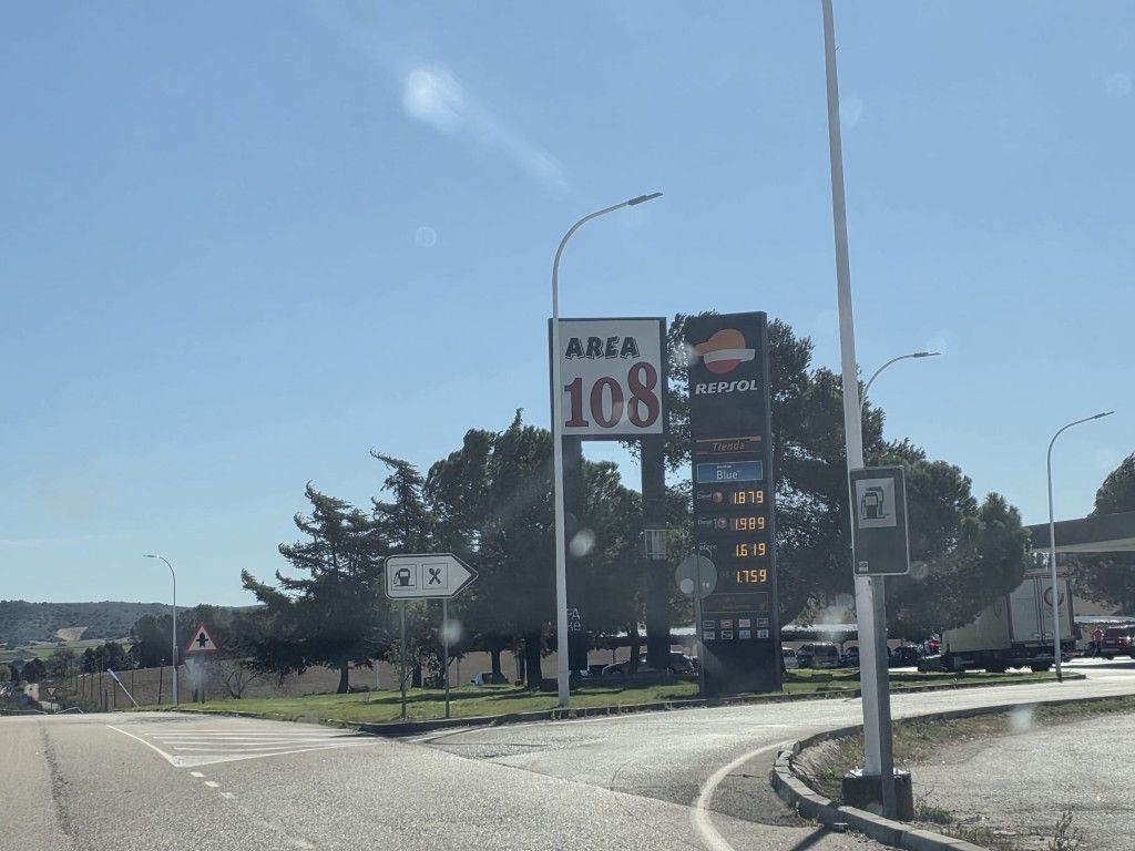 Señal de la zona 108 al borde de la carretera bajo un cielo azul despejado, con árboles y un aparcamiento cerca.