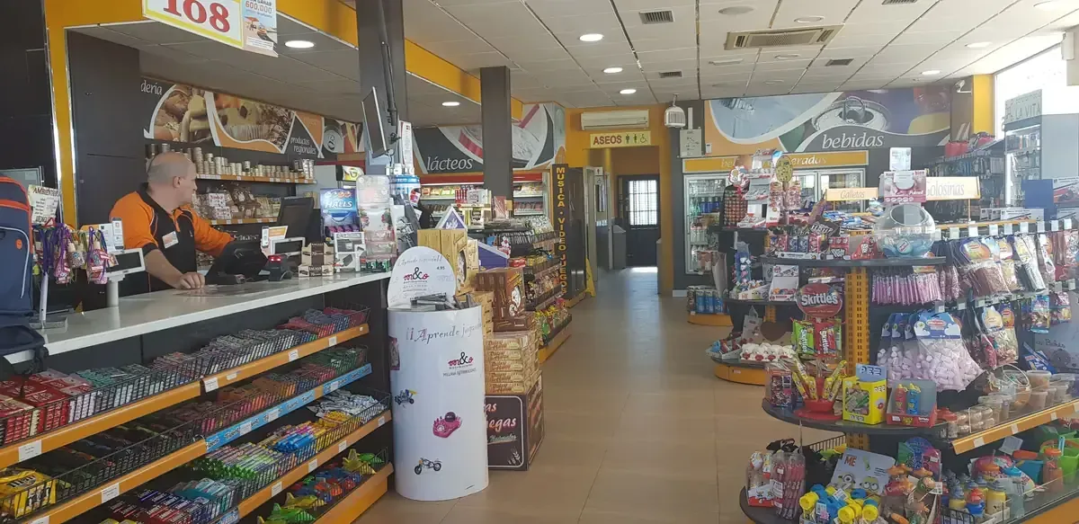 Pasillo de tienda de conveniencia con bocadillos, bebidas y un mostrador que exhibe mercancía variada.