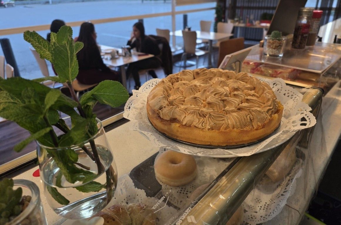 Un pastel de manzana sobre el mostrador de una cafetería luminosa, con una pequeña planta y gente sentada junto a la ventana.