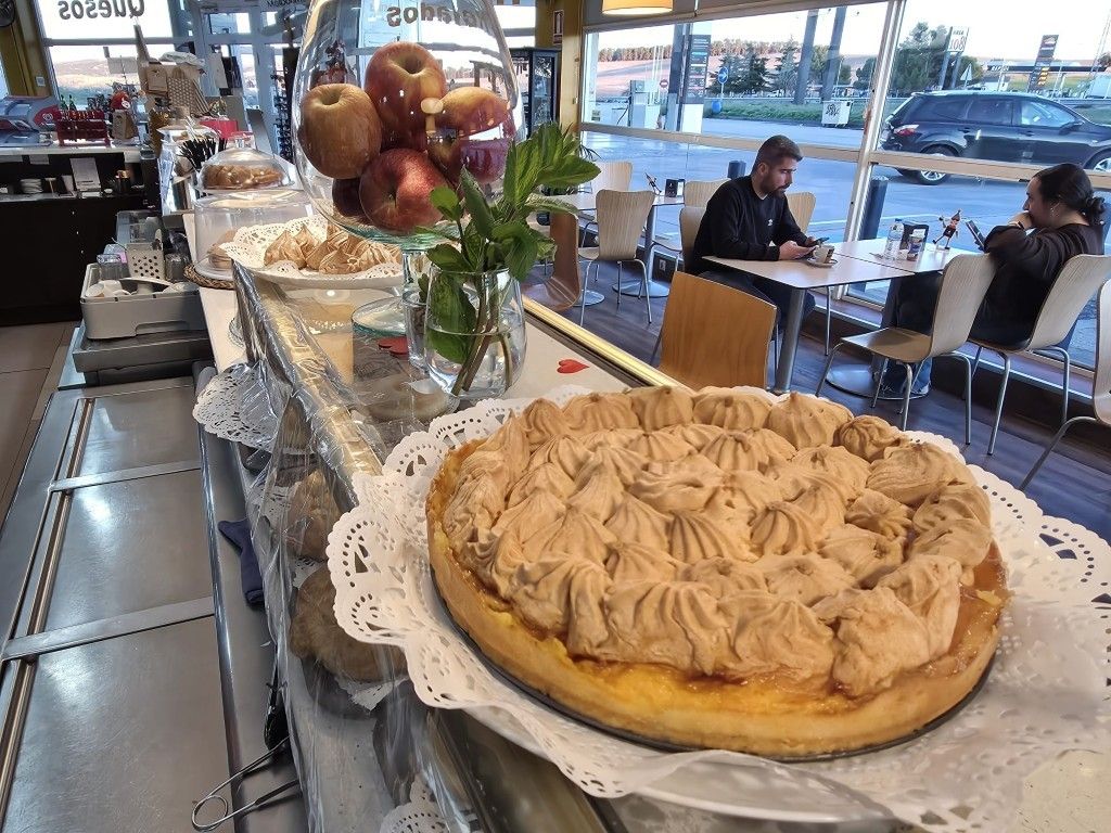 Mostrador de cafetería con una gran tarta cubierta de crema en un plato y gente sentada junto a la ventana.