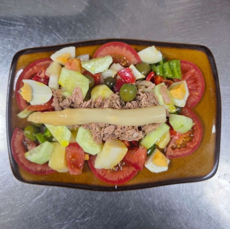 Ensalada con atún, huevos duros, aceitunas, tomates y pepino en un plato rectangular marrón.