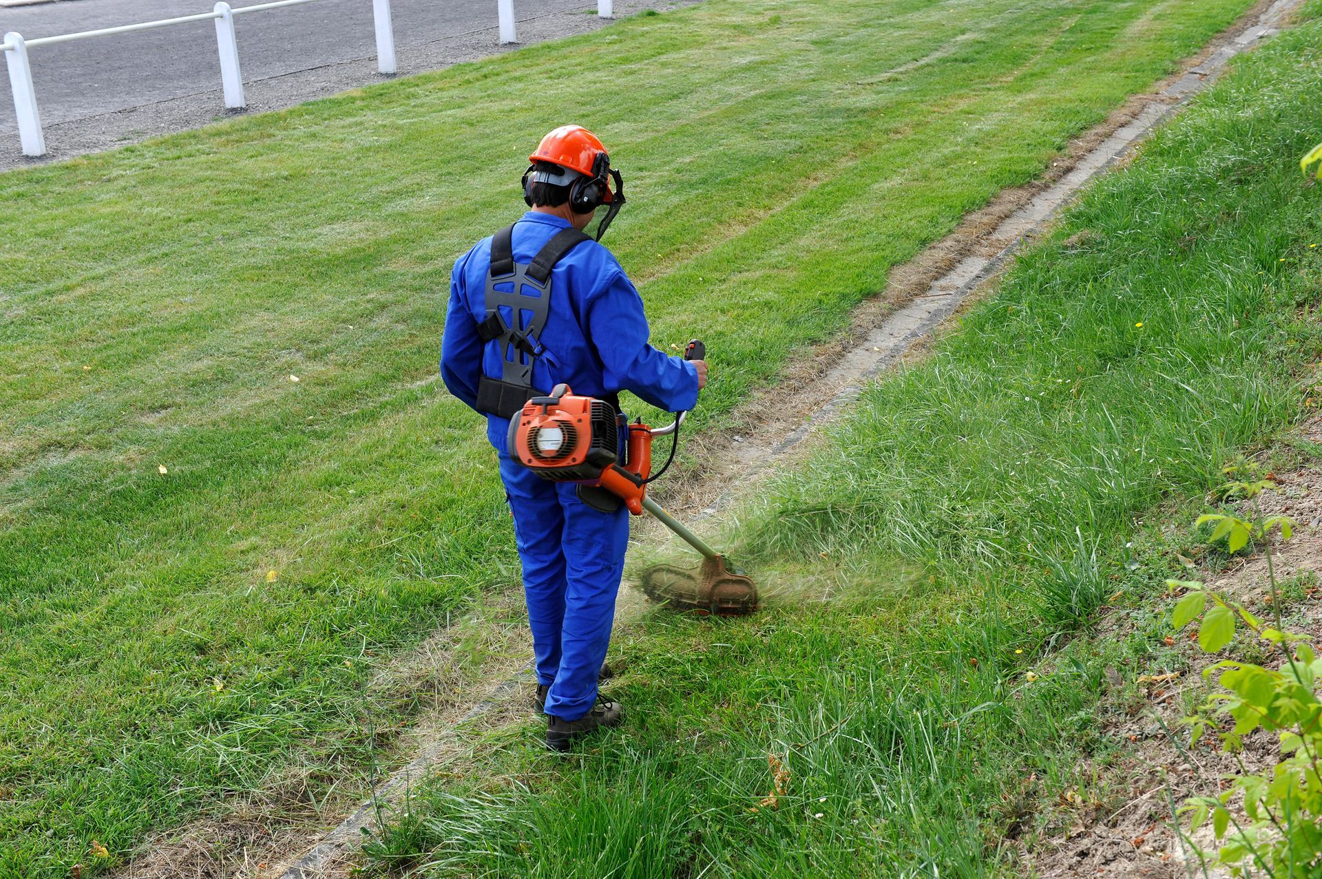 Un ouvrier en uniforme bleu coupe l'herbe avec un coupe-bordure sur le bord d'une route en pente.