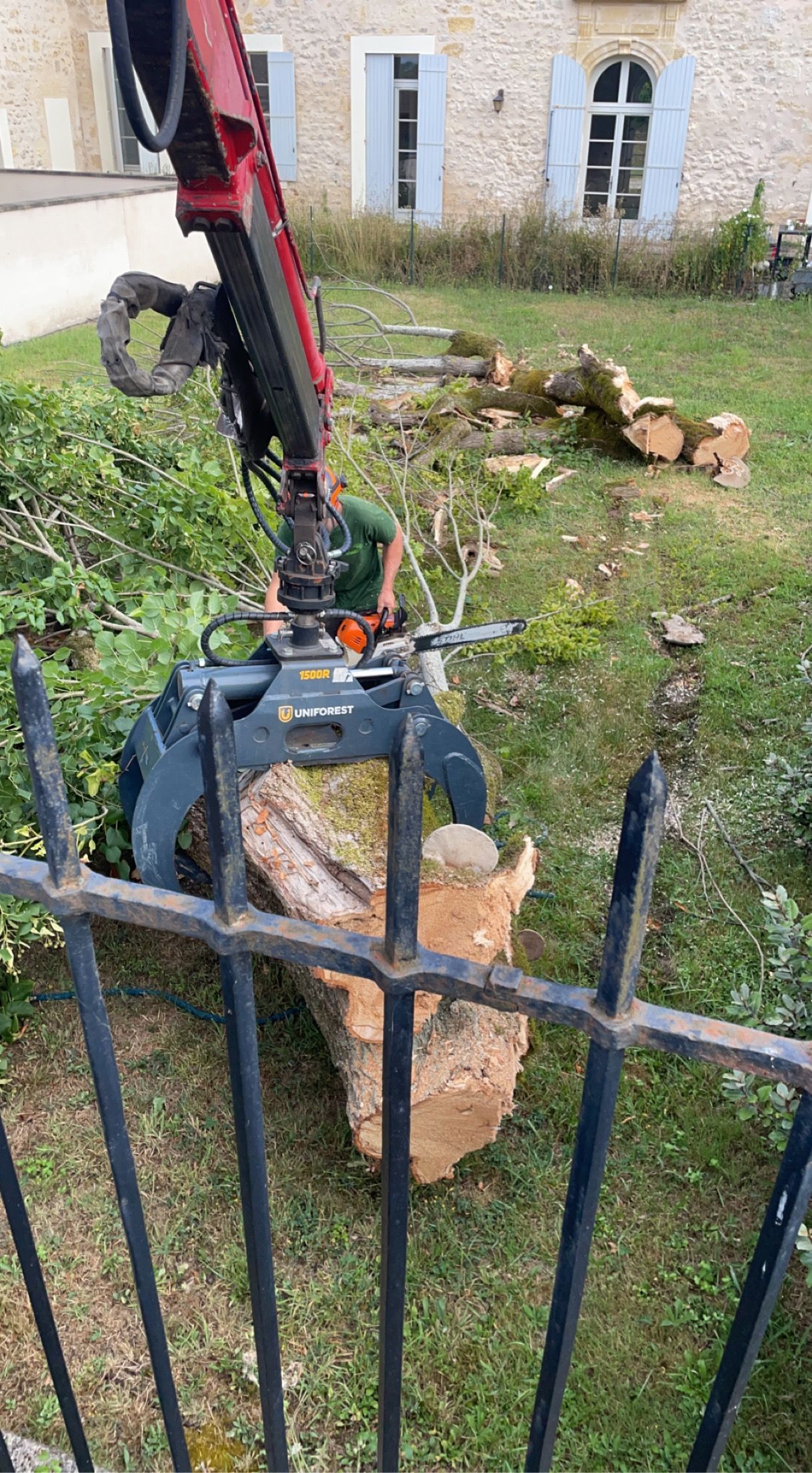 Un bras d'excavatrice rouge soulève une grosse souche d'arbre à côté d'une clôture métallique noire dans une cour avec de l'herbe.
