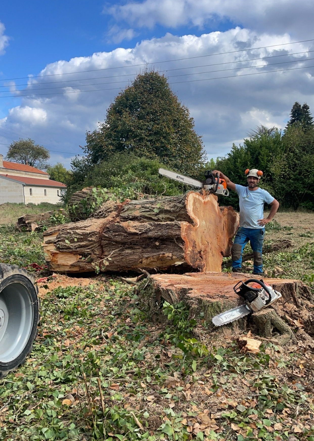 Deux personnes posent à côté d'une souche d'arbre fraîchement coupée et d'une tronçonneuse dans une cour avec de l'herbe sous un ciel bleu.