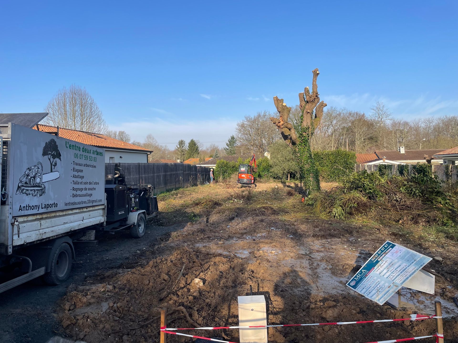 Chantier de construction au sol boueux, un camion à gauche et des ouvriers déblayant les débris sous un ciel bleu