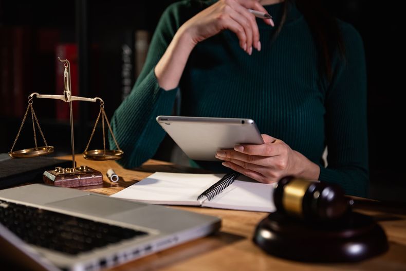 Photo d'une femme avec une tablette, un mateau d'avocat devant elle et la balance de la justice.