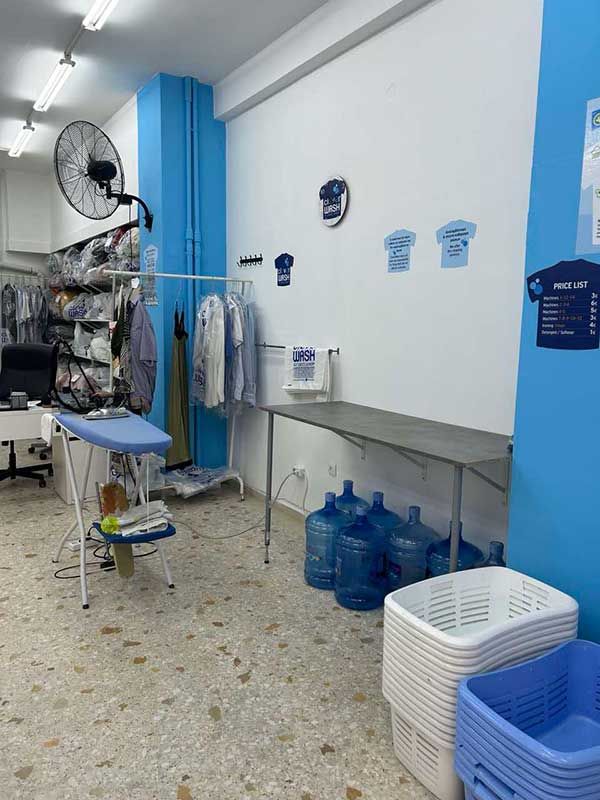 Ένα πλυσταριό με τραπέζι, σιδερώστρα, καλάθια και μπουκάλια νερό.