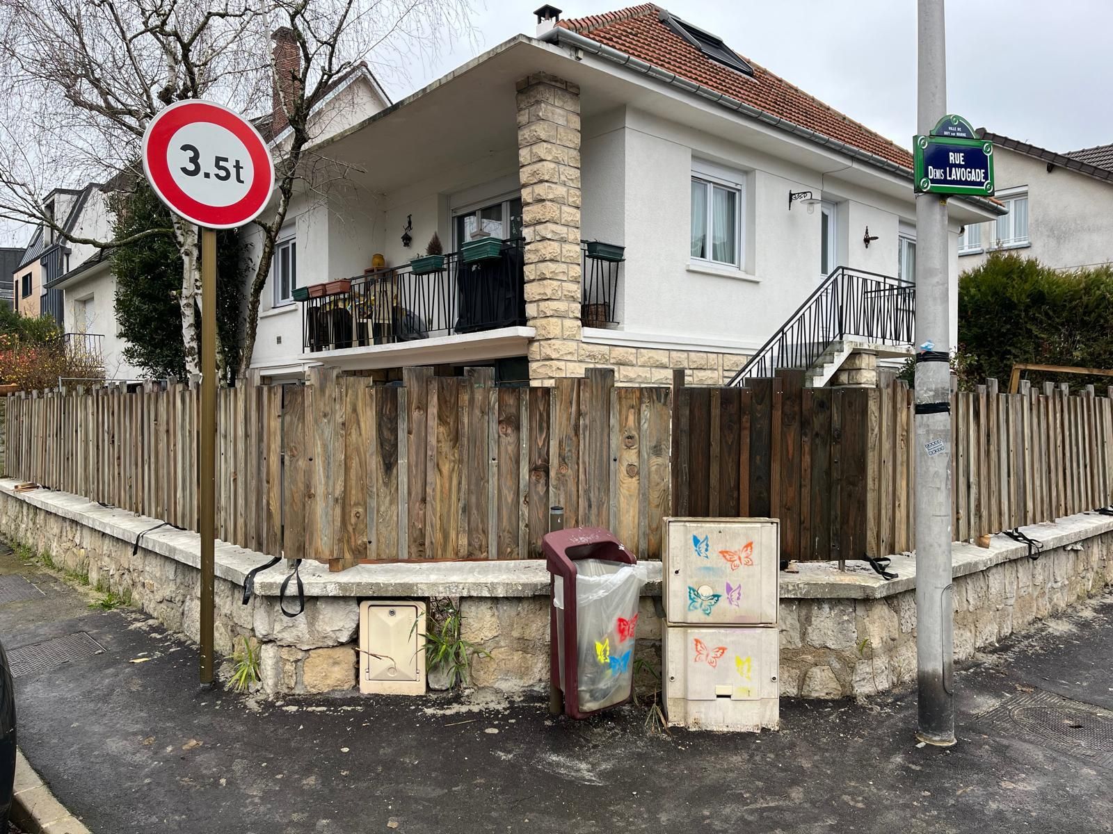Maison d'angle résidentielle derrière une clôture en bois, avec des poubelles et un panneau de signalisation.