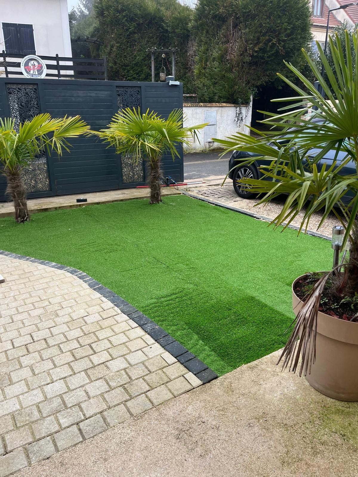 Petite cour paysagée avec gazon artificiel, pavés, palmiers et une voiture garée en arrière-plan