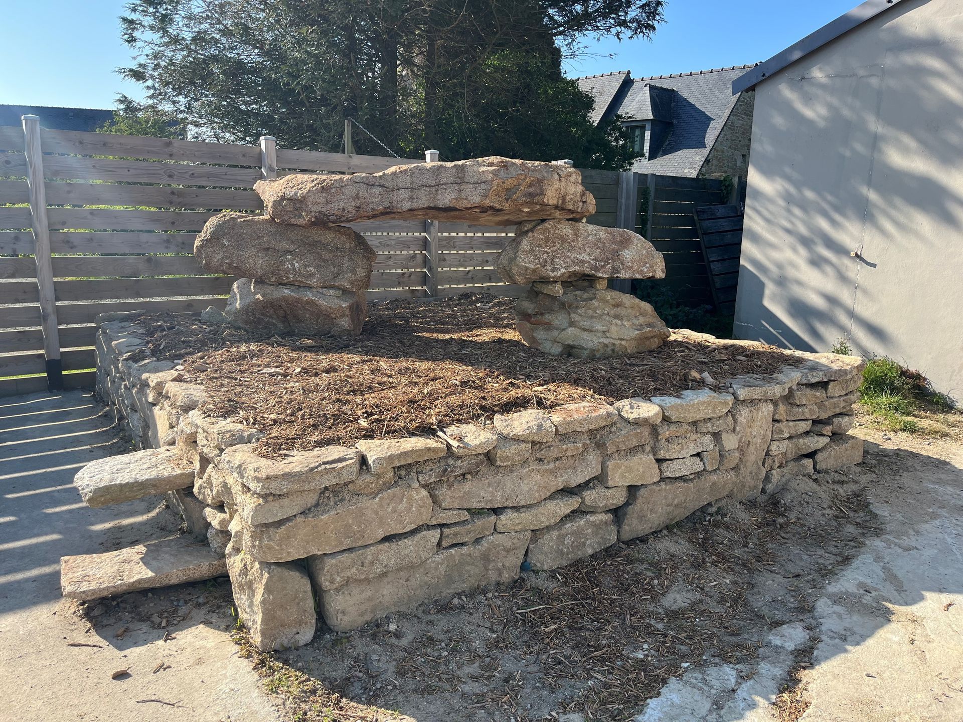 Fontaine en pierres empilées dans un jardin ensoleillé, à côté d'une clôture et d'une maison, entourée d'un sol gravillonné.