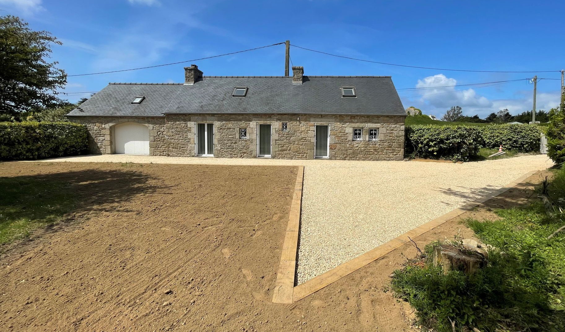 Maisonnette en pierre au toit gris, précédée d'une allée de gravier et de terre sous un ciel d'un bleu éclatant.