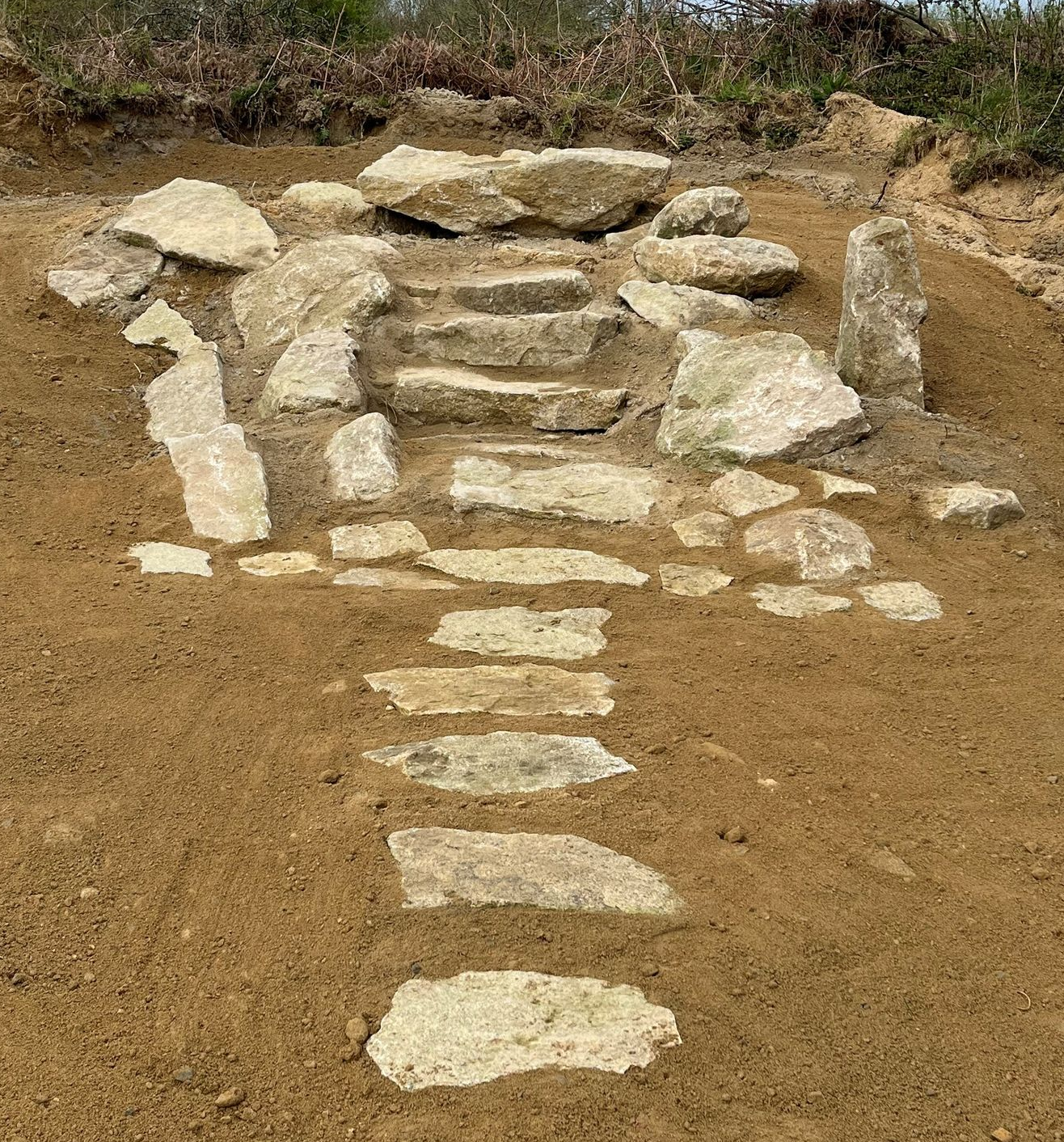 Des marches en pierre aménagées dans une pente de terre avec des pierres plates mènent à une zone herbeuse.