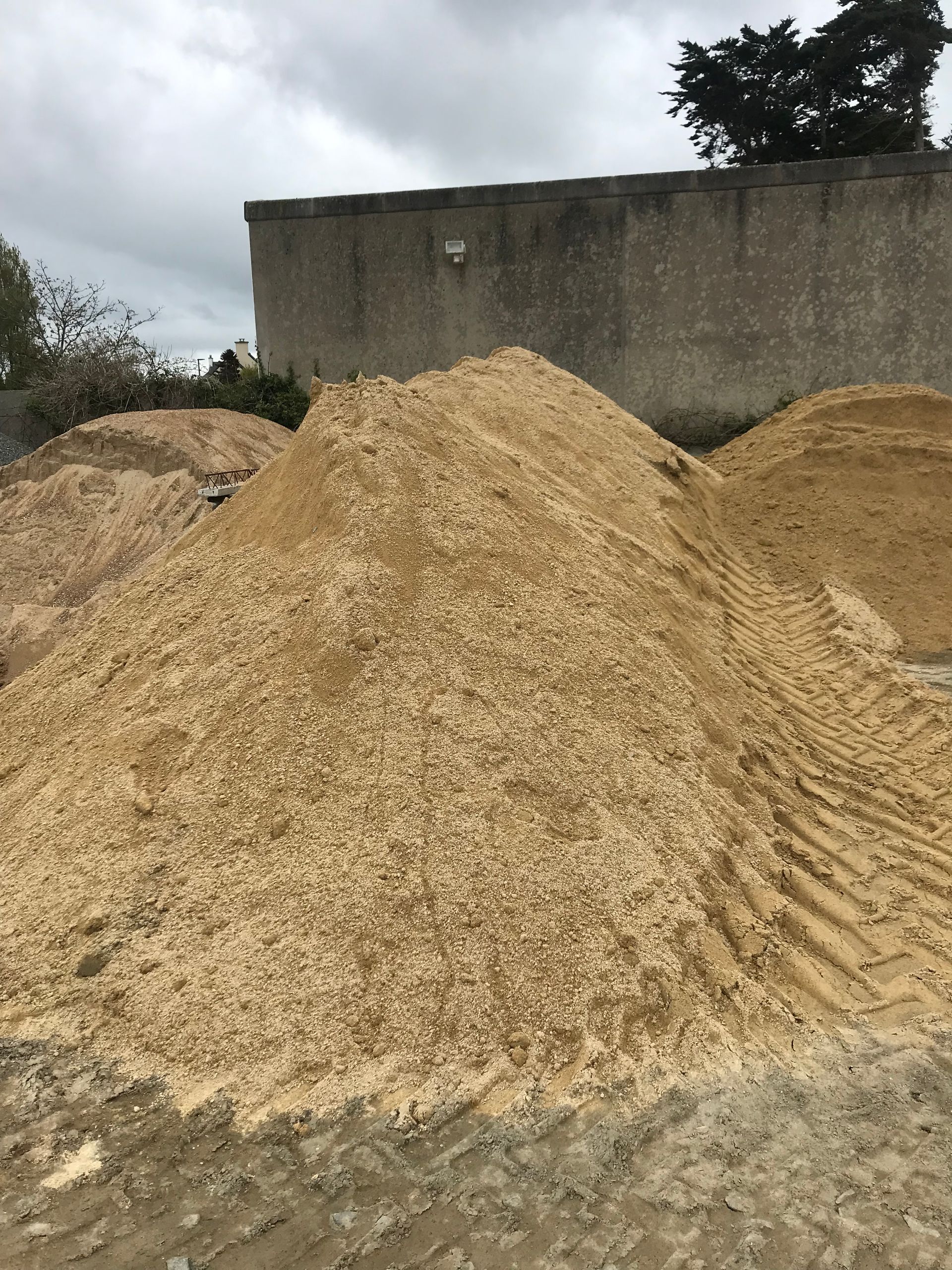 Gros tas de sciure ou de copeaux de bois à l'extérieur, à côté d'un mur en béton, sous un ciel nuageux.