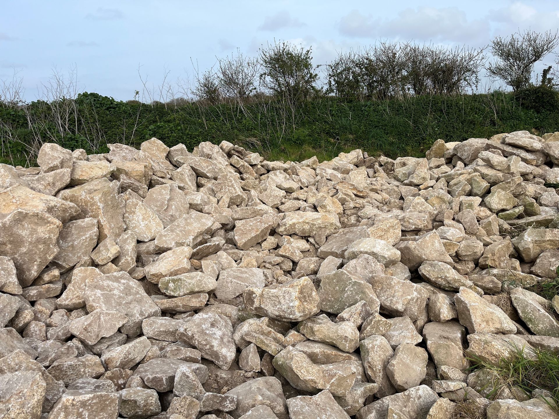 Des rochers beiges empilés au premier plan, des arbustes verts et un ciel nuageux en arrière-plan.