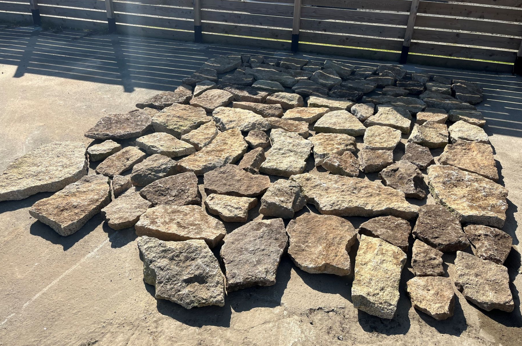 Un tas de dalles de pierre brute disposées sur du béton au soleil, avec une clôture métallique en arrière-plan.