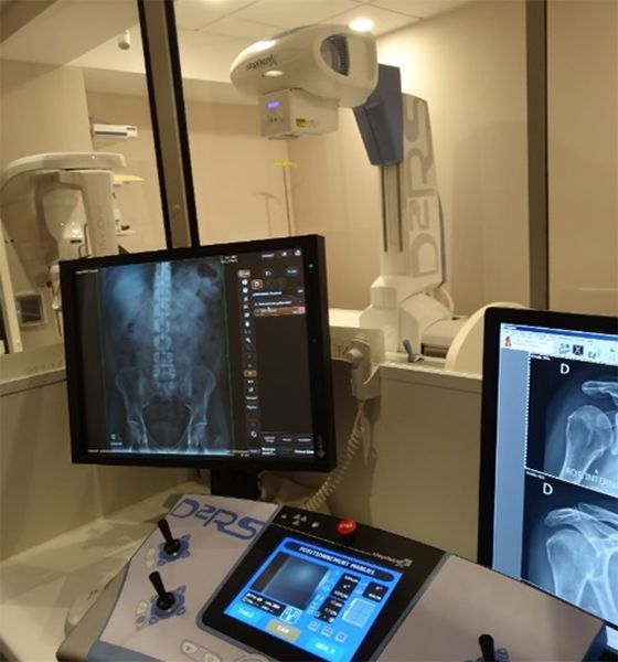 Salle de radiologie équipée d'un appareil de radiographie vertical, d'un moniteur patient et d'écrans de console affichant les examens.