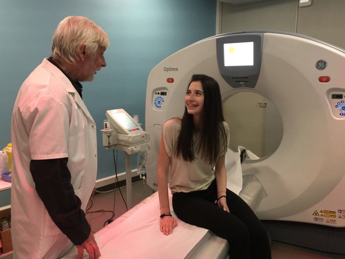 Une femme assise à côté d'un scanner médical discute avec un homme en chemise rose dans une clinique.
