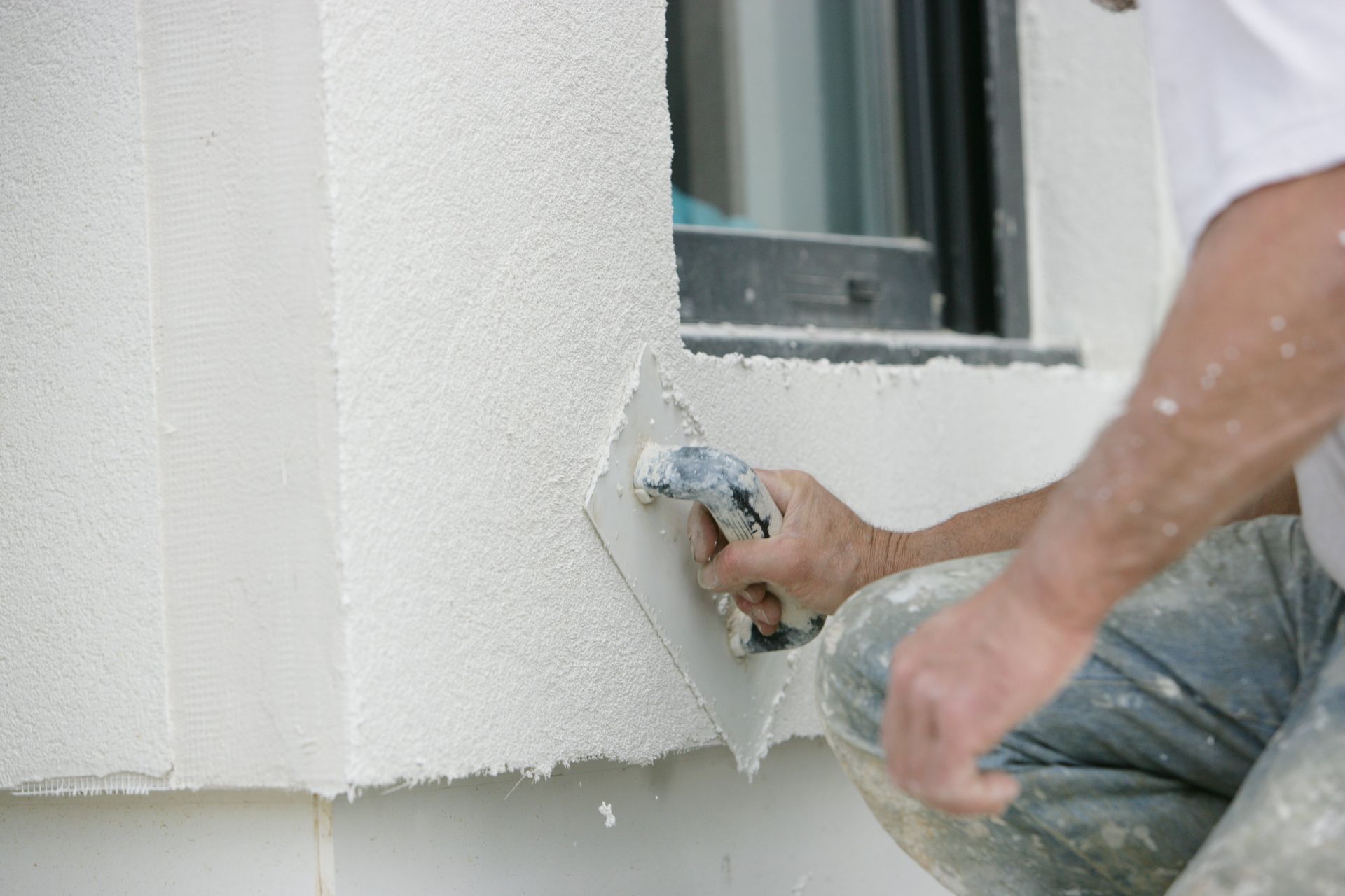 Personne appliquant du crépis avec une truelle sur un mur extérieur près d'une fenêtre.