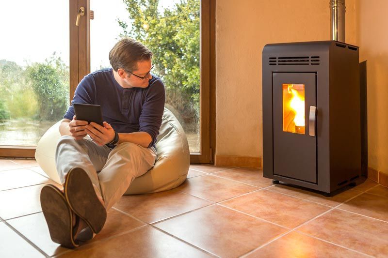 Homme assis près de la fenêtre avec une tablette, près d'un poêle à granulés avec des flammes.