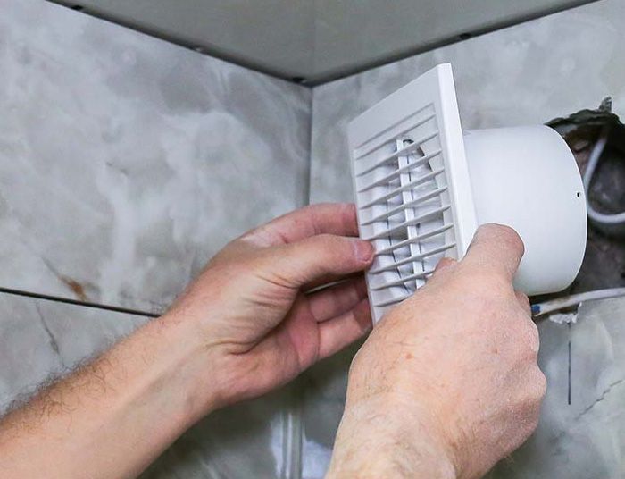 Personne installant un ventilateur d'extraction blanc dans une salle de bain sur un mur carrelé.