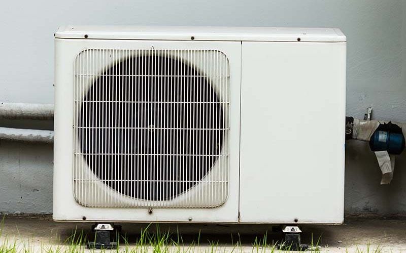 Unité de climatisation extérieure blanche, avec grille de ventilation et panneau latéral, montée sur blocs noirs.