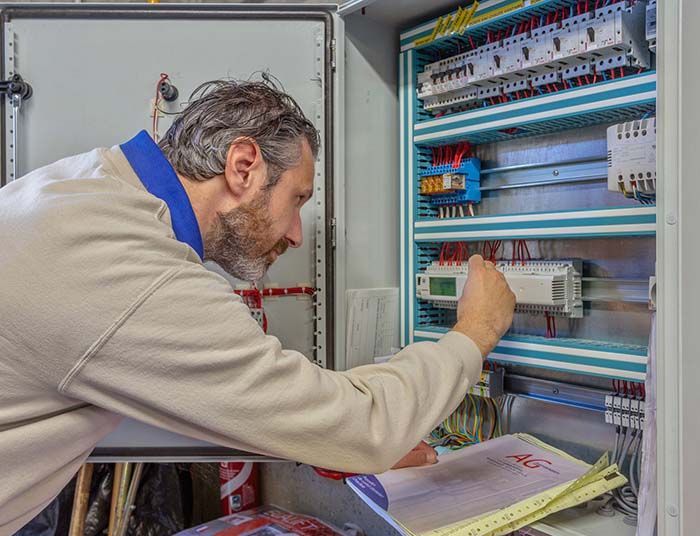 Un homme en pull clair travaille sur un tableau électrique, des documents à la main.